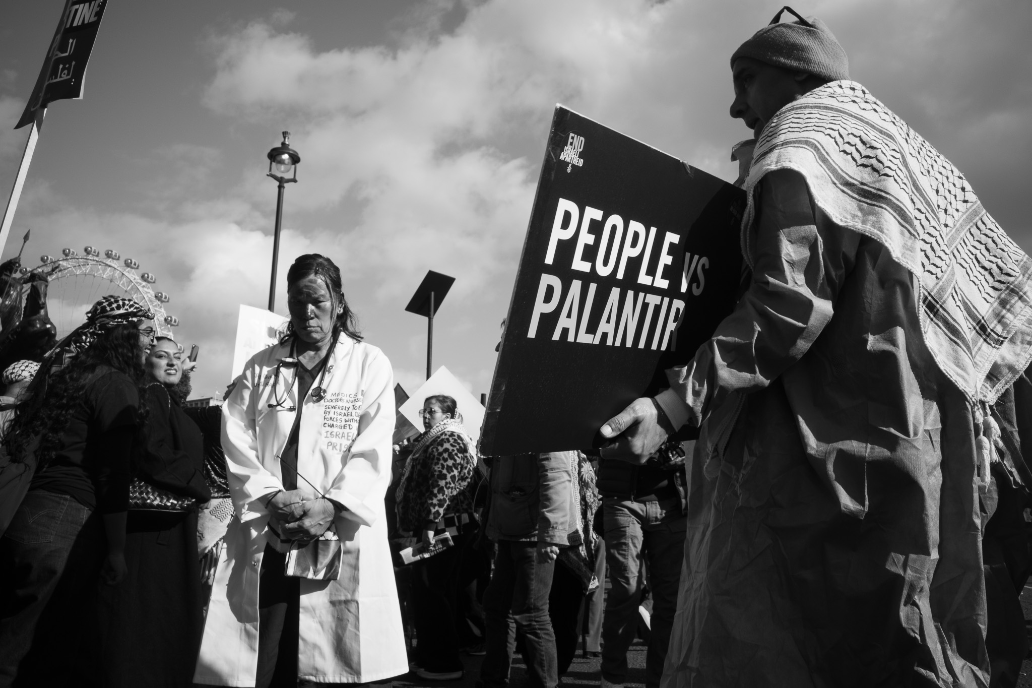 A woman in a white coat with a stethoscope and a marked face standing still among marchers, a black placard in the foreground