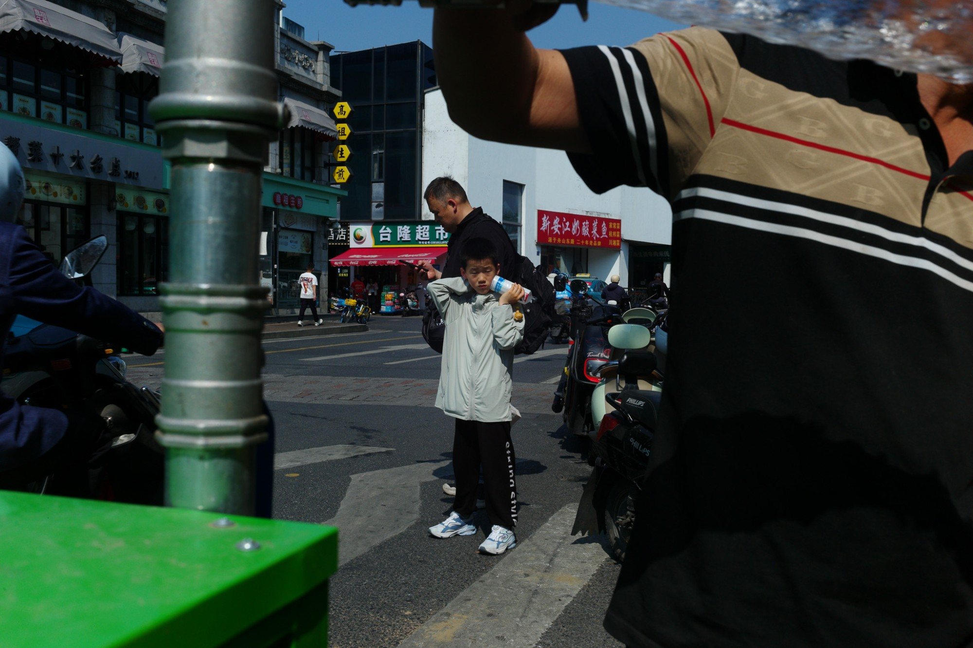 A boy holding a bottle to his ear at a crossing, framed between a pole and a passer-by's arm