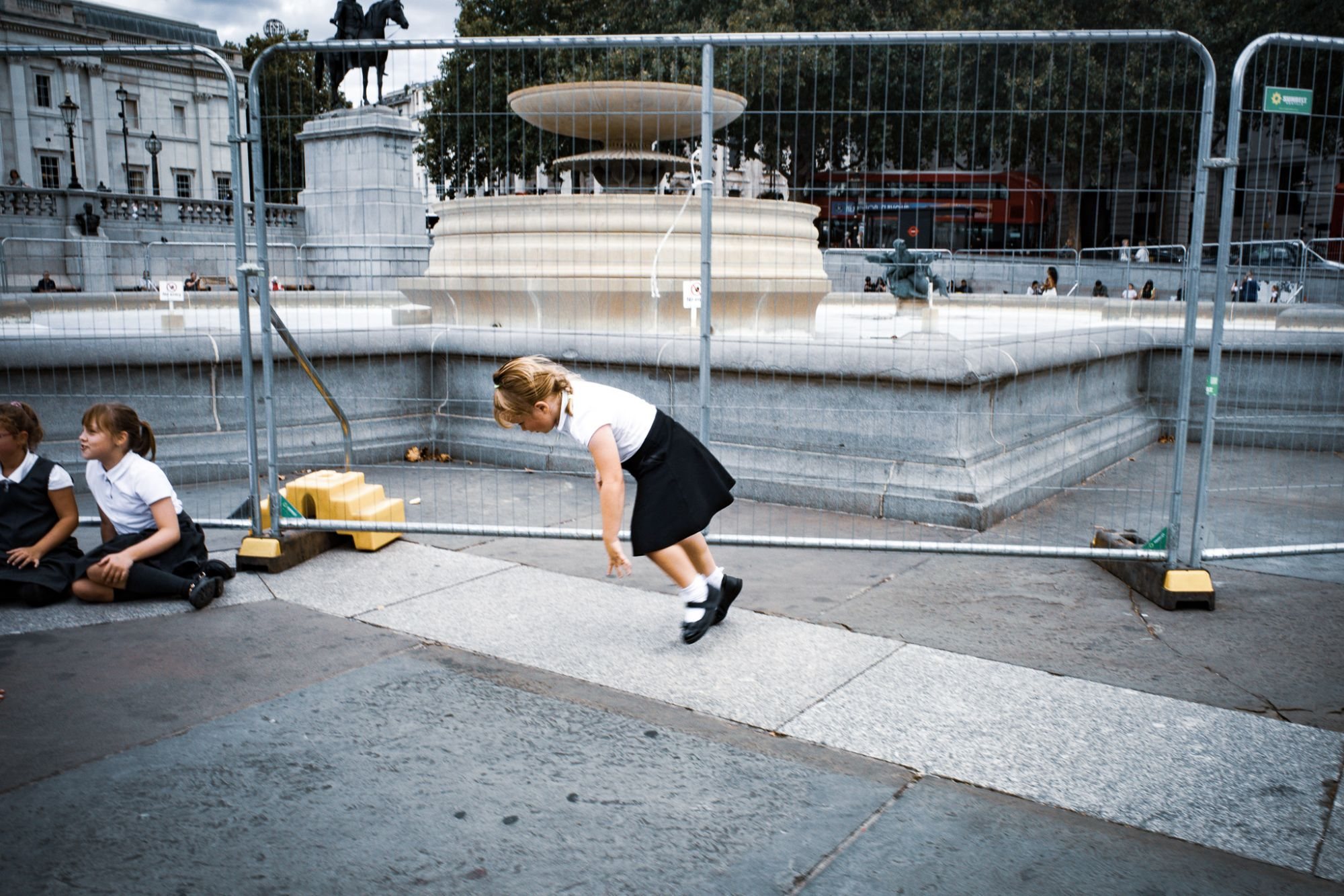 A schoolgirl leaning mid-step beside a fenced-off fountain in Trafalgar Square