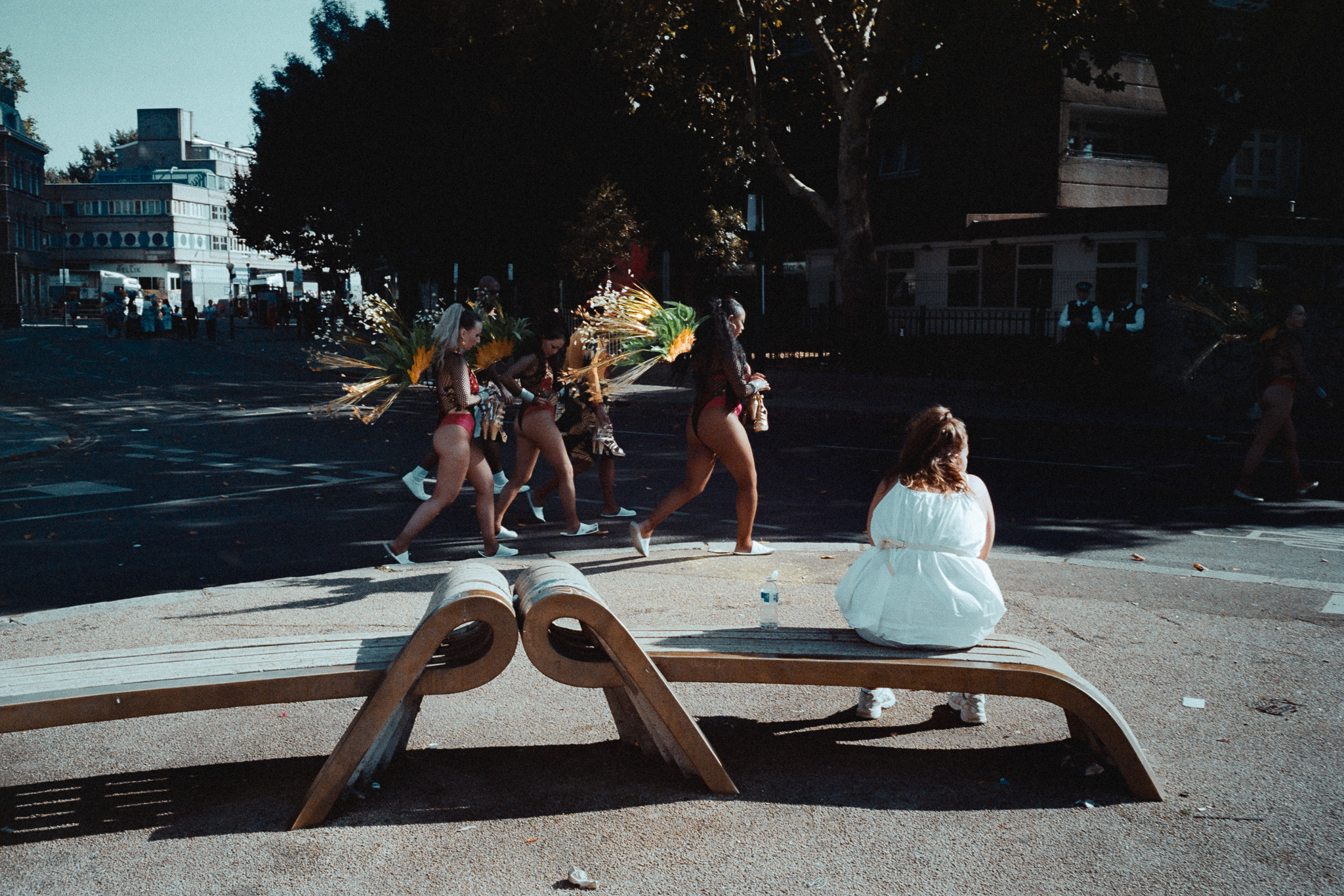 Carnival performers walking away down a road while a woman in white sits on a bench